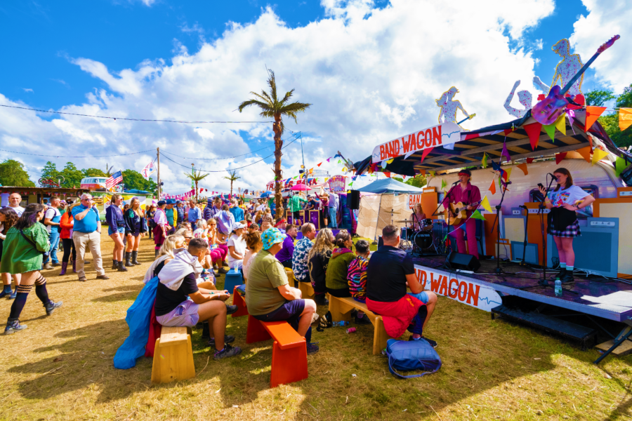 Blue skies with some fluffy white clouds, People sitting down watching a performance at the Band Wagon stage at Trailer Park, Electric Picnic.